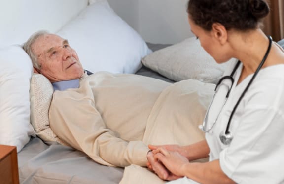 Elderly man laying in bed with a caregiver sitting at the edge of the bed